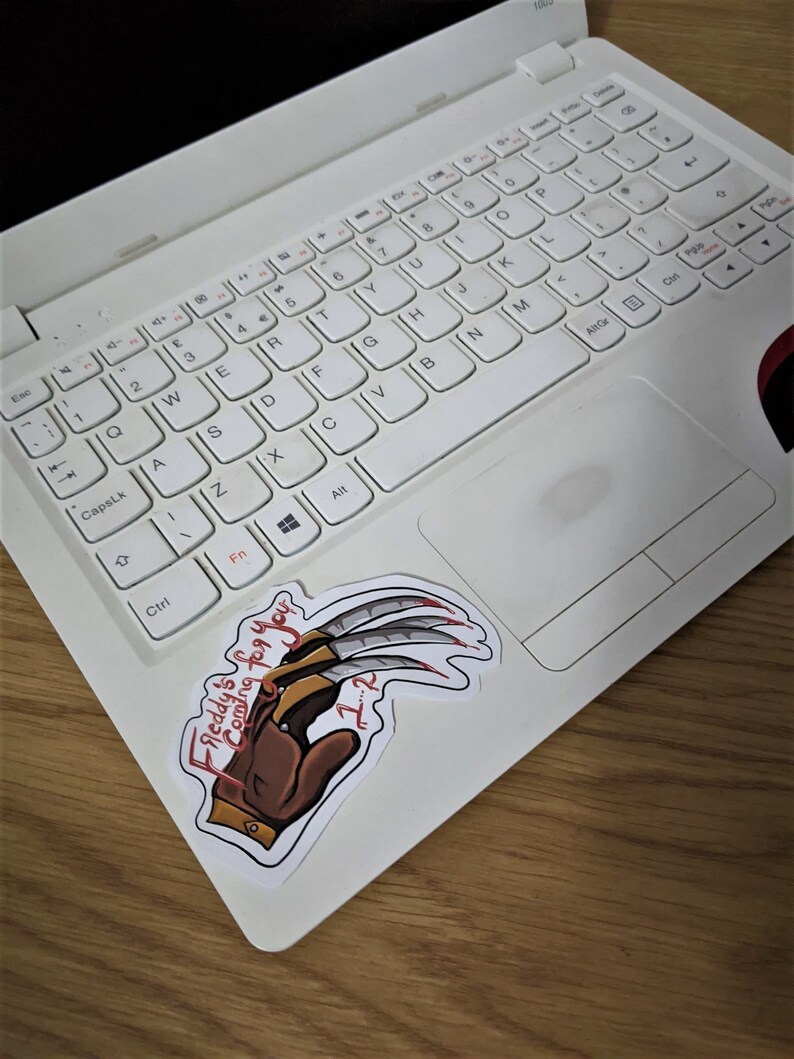 K&ouml;nnte beinhalten: Ein wei&szlig;er Laptop mit einem Freddy Krueger-Aufkleber auf der rechten Seite des Touchpads. Der Aufkleber zeigt eine Zeichnung von Freddys Handschuh mit den Worten "Freddy's is coming for you" und "7-2" darauf.