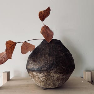 May include: A handcrafted, dark brown ceramic vase with a textured surface. The vase is adorned with a branch of dried leaves. The vase is displayed on a light-coloured wooden shelf, creating a natural, earthy aesthetic.