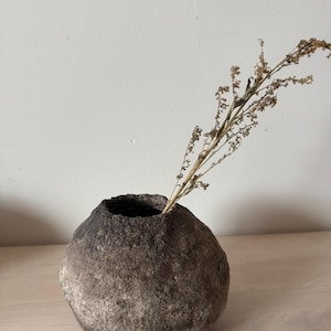 Könnte beinhalten: Eine runde, strukturierte, graue Vase mit dunkler Öffnung, die einen getrockneten, braunen Pflanzenstiel enthält. Die Vase hat ein raues, steinartiges Aussehen und steht auf einer hellen Holzoberfläche.