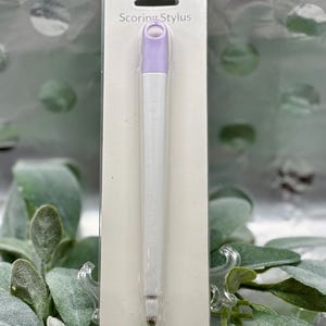 May include: A white and lavender Scoring Stylus in a clear plastic package. The stylus has a lavender top and a white body. The text "Scoring Stylus" is printed on the package. The stylus is designed for crafting and paper projects.