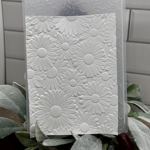 Op de afbeelding: Een rechthoekige witte embossing folder met een verhoogd, getextureerd patroon van meerdere madeliefjes met gedetailleerde harten en bloemblaadjes. De doorzichtige plastic map is rechtop geplaatst en toont een bloemmotief.