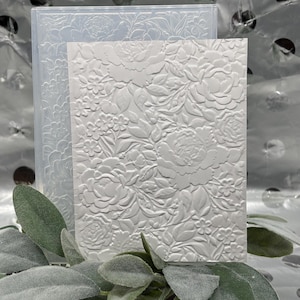 Op de afbeelding: Een witte, in reliëf gemaakte bloemenontwerp op een rechthoekige kaart en een doorzichtige plastic embossing folder. Het ontwerp toont gedetailleerde bloemen en bladeren. De kaart is voor de map geplaatst.
