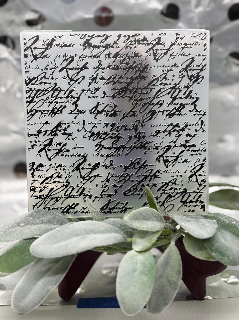 May include: A rectangular, frosted glass panel with black script writing. The panel is positioned in front of a sprig of sage leaves with a burgundy base. The script appears to be an old handwritten letter.