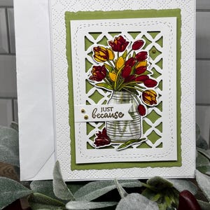 May include: A handmade greeting card featuring a white die-cut lattice frame over a sage green background. The center displays a bouquet of red and yellow tulips in a glass jar with a white banner printed with the text "JUST because" in black.