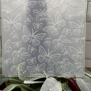 May include: A square, clear plastic embossing folder featuring a repeating pattern of various sized butterflies with detailed wing veins. The design is etched into the surface, creating a raised texture intended for paper crafting and card making.
