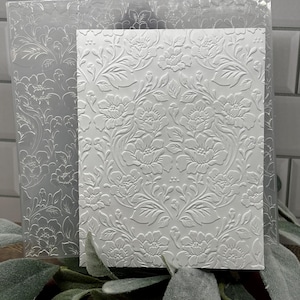 Könnte beinhalten: Ein transparenter Prägefolder aus Kunststoff mit einem detaillierten floralen Damastmuster, kombiniert mit einem weißen Kartonblatt, das dasselbe Blumen- und Blattdesign als erhabenes, texturiertes Relief auf der gesamten Oberfläche zeigt.