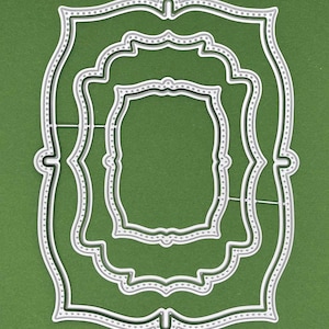 May include: A set of three nested silver metal cutting dies, each featuring a decorative scalloped frame shape with a dotted border pattern. The dies are arranged concentrically, with the largest on the outside and the smallest in the center.
