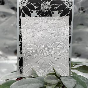 May include: A white embossed sunflower design on a rectangular card. A black and white sunflower design is visible on the side. The card is placed on a bed of green leaves.