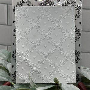 Op de afbeelding: Een rechthoekige witte embossing folder met een verhoogd ruitpatroon bestaande uit delicate bloemenranken en kleine hartjes. Het ontwerp creëert een gestructureerd, herhalend motief over het oppervlak, geschikt voor papierknutselprojecten.