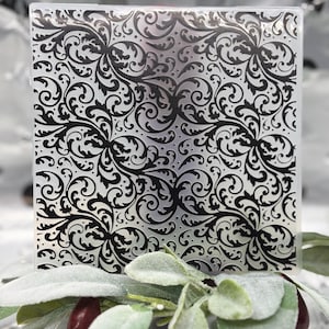 May include: A clear, square decorative block with a black, swirling floral pattern. The design is intricate and detailed, with a vintage aesthetic. The block is set against a blurred background with greenery and berries.