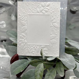May include: A white embossed card with a rectangular frame and floral and leaf designs. A clear, textured plastic mold is behind the card. The card is resting on a bed of green leaves and a dark red accent.