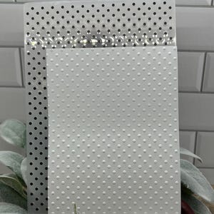 Op de afbeelding: Een rechthoekige doorzichtige plastic embossing folder met een herhalend patroon van kleine, gelijkmatig verdeelde cirkelvormige inkepingen, geplaatst over een wit vel karton dat dezelfde verhoogde stippentextuur over het gehele oppervlak toont.