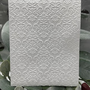 Może przedstawiać: Biała, prostokątna karta z wytłoczonym wzorem damasceńskim. Złożony wzór przedstawia powtarzające się motywy kwiatowe i zwoje, tworząc teksturowaną powierzchnię. Karta otoczona jest zielonymi liśćmi i ciemnoczerwonym akcentem.