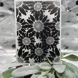 K&ouml;nnte beinhalten: Ein rechteckiger, klarer Glasgegenstand mit einem schwarzen Sonnenblumenmuster. Die Sonnenblumen sind in einem sich wiederholenden Muster angeordnet. Das Objekt liegt auf gr&uuml;nen und roten Bl&auml;ttern.