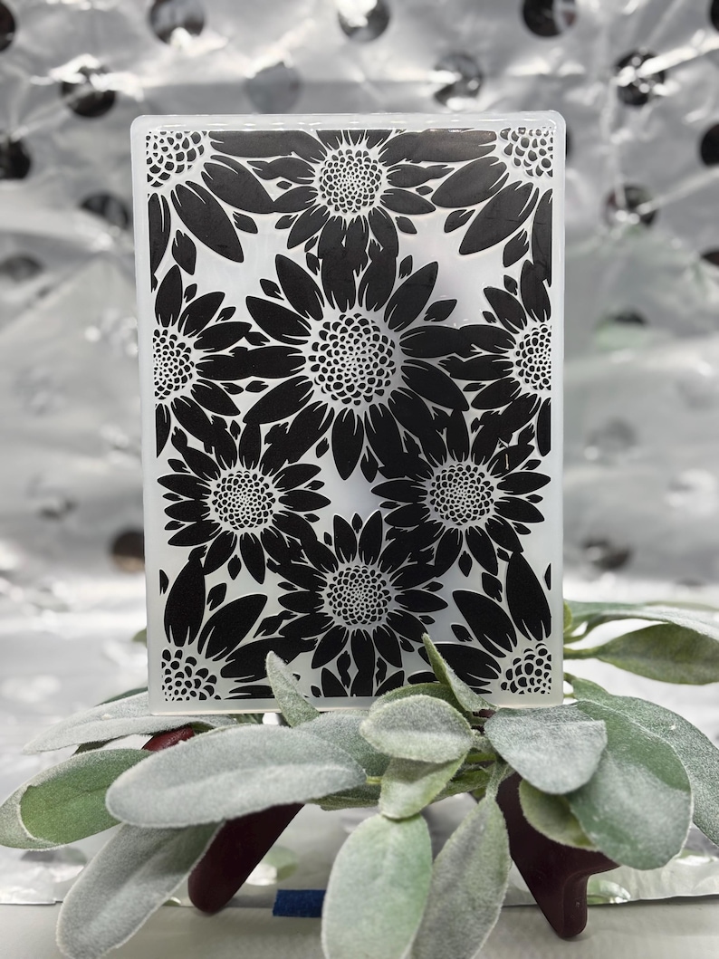 K&ouml;nnte beinhalten: Ein rechteckiger, klarer Glasgegenstand mit einem schwarzen Sonnenblumenmuster. Die Sonnenblumen sind in einem sich wiederholenden Muster angeordnet. Das Objekt liegt auf gr&uuml;nen und roten Bl&auml;ttern.