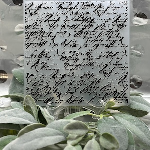 May include: A square, clear glass tile with black script writing. The tile is displayed on a clear stand, surrounded by green and gray foliage. The background is a silver, polka-dotted surface.