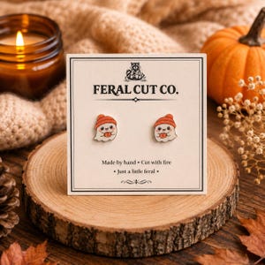 May include: A pair of small stud earrings featuring a white ghost character wearing an orange beanie and holding a mug. The earrings are attached to a square display card labeled FERAL CUT CO. with the text Made by hand, Cut with fire, Just a little feral.