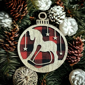 May include: A wooden Christmas ornament shaped like a round ball with a red and black buffalo plaid pattern. The ornament has a cutout of a rocking horse inside the plaid pattern.