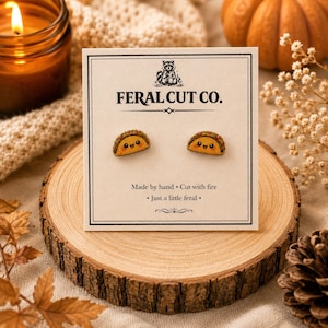 May include: A pair of small, wooden taco-shaped stud earrings with smiling faces, displayed on a white card labeled FERAL CUT CO. The card features a raccoon illustration and the text Made by hand, Cut with fire, and Just a little feral.