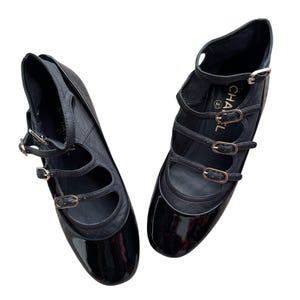 May include: Pair of black leather Mary Jane style flats with multiple straps and gold-tone buckles. The shoes have a glossy patent leather toe and a matte leather upper. The design includes a small heel and a strap around the ankle.