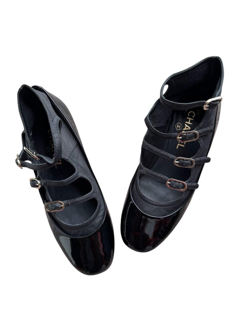 May include: Pair of black leather Mary Jane style flats with multiple straps and gold-tone buckles. The shoes have a glossy patent leather toe and a matte leather upper. The design includes a small heel and a strap around the ankle.