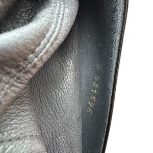 May include: Close-up of black leather material with visible stitching and a shiny, smooth surface. Gold lettering reads "965196 G" on the right side. The leather appears to be part of a shoe or bag.