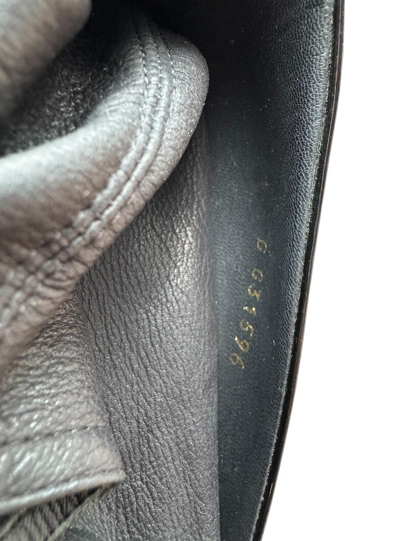 May include: Close-up of black leather material with visible stitching and a shiny, smooth surface. Gold lettering reads "965196 G" on the right side. The leather appears to be part of a shoe or bag.