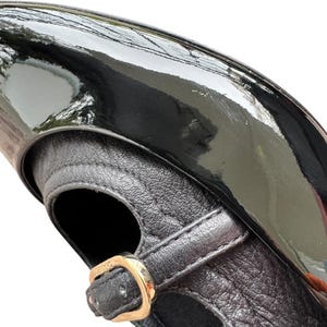 May include: Close-up of a black patent leather shoe with a strap and gold-colored buckle. The shoe has a glossy finish and a textured leather strap. The design is classic and elegant.