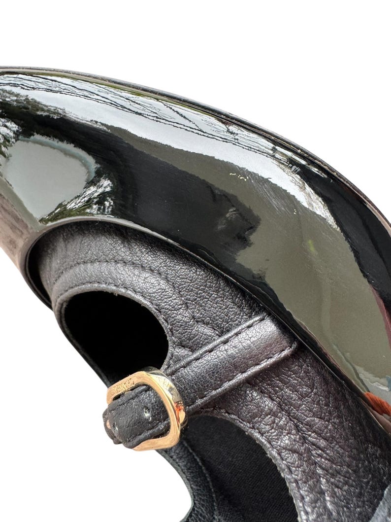May include: Close-up of a black patent leather shoe with a strap and gold-colored buckle. The shoe has a glossy finish and a textured leather strap. The design is classic and elegant.
