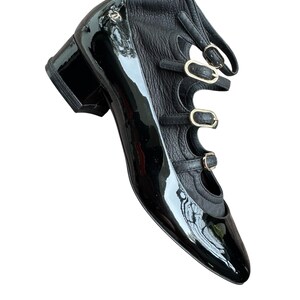 May include: Black patent leather shoe with a pointed toe and a block heel. The shoe features three straps with gold-tone buckles and a cutout design. A small logo is visible on the side.