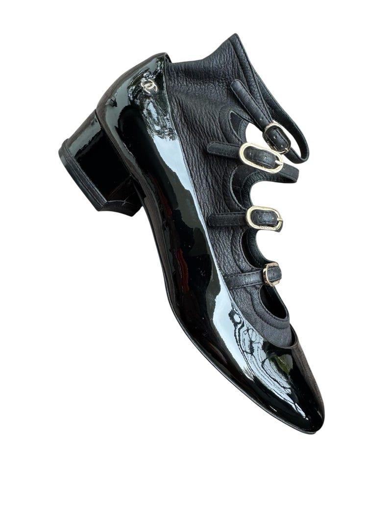 May include: Black patent leather shoe with a pointed toe and a block heel. The shoe features three straps with gold-tone buckles and a cutout design. A small logo is visible on the side.