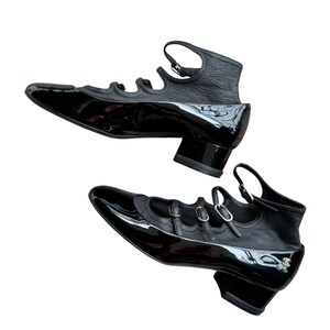 May include: Pair of black patent leather shoes with a pointed toe and ankle strap. The shoes have a low block heel and multiple straps across the top. The design includes a textured leather section at the back of the shoe.