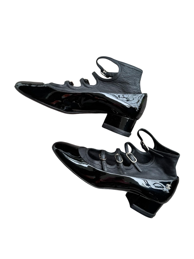 May include: Pair of black patent leather shoes with a pointed toe and ankle strap. The shoes have a low block heel and multiple straps across the top. The design includes a textured leather section at the back of the shoe.