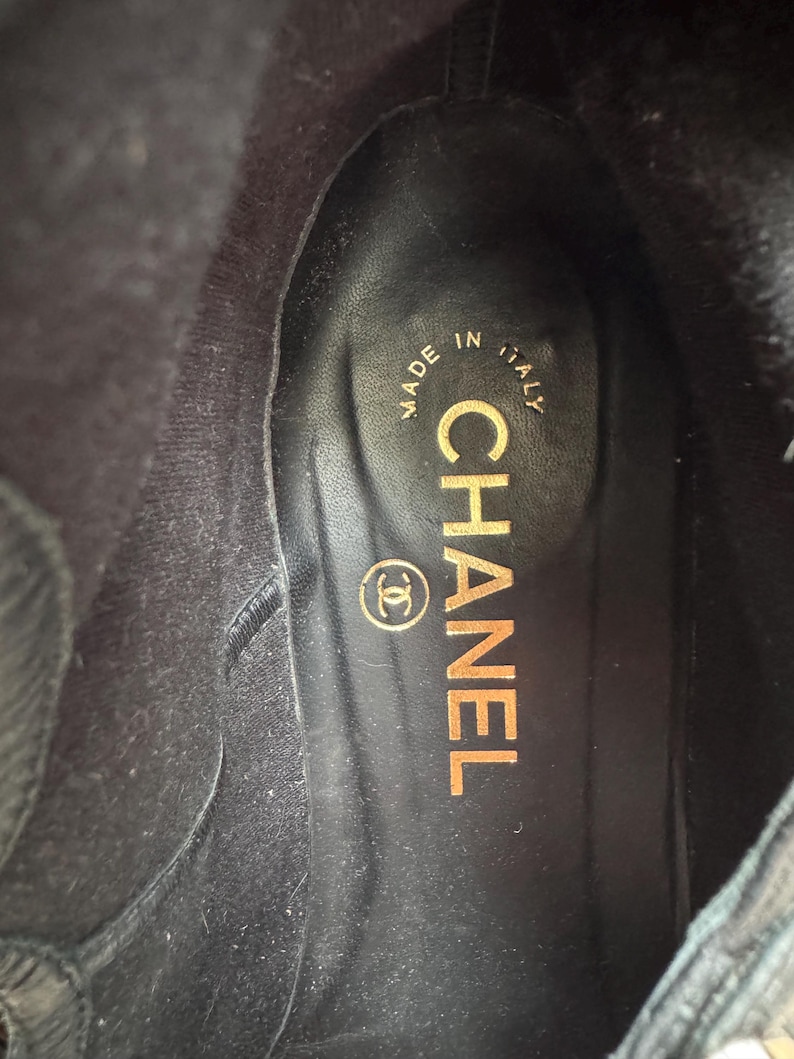 May include: Interior view of a black boot with a leather insole. The insole features the gold-colored text "CHANEL" and the Chanel logo. The text "MADE IN ITALY" is also visible.