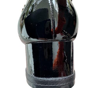 May include: Close-up of a black patent leather shoe with a block heel. The shoe has a glossy finish and a rounded toe. The heel is approximately 2 inches high.