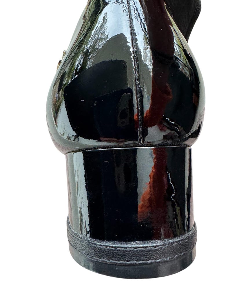 May include: Close-up of a black patent leather shoe with a block heel. The shoe has a glossy finish and a rounded toe. The heel is approximately 2 inches high.