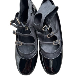 May include: Pair of black leather and patent leather shoes with multiple straps and gold-tone buckles. The shoes have a Mary Jane style with a high vamp and ankle strap. The brand name is visible on the shoe.