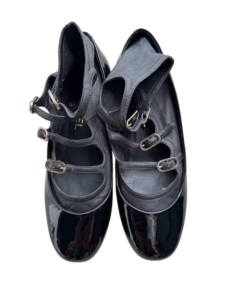 May include: Pair of black leather and patent leather shoes with multiple straps and gold-tone buckles. The shoes have a Mary Jane style with a high vamp and ankle strap. The brand name is visible on the shoe.