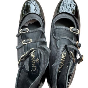 May include: Pair of black leather Mary Jane style shoes with patent leather toe caps. The shoes have multiple straps with gold-tone buckles and the brand name "CHANEL" is visible inside the shoe.
