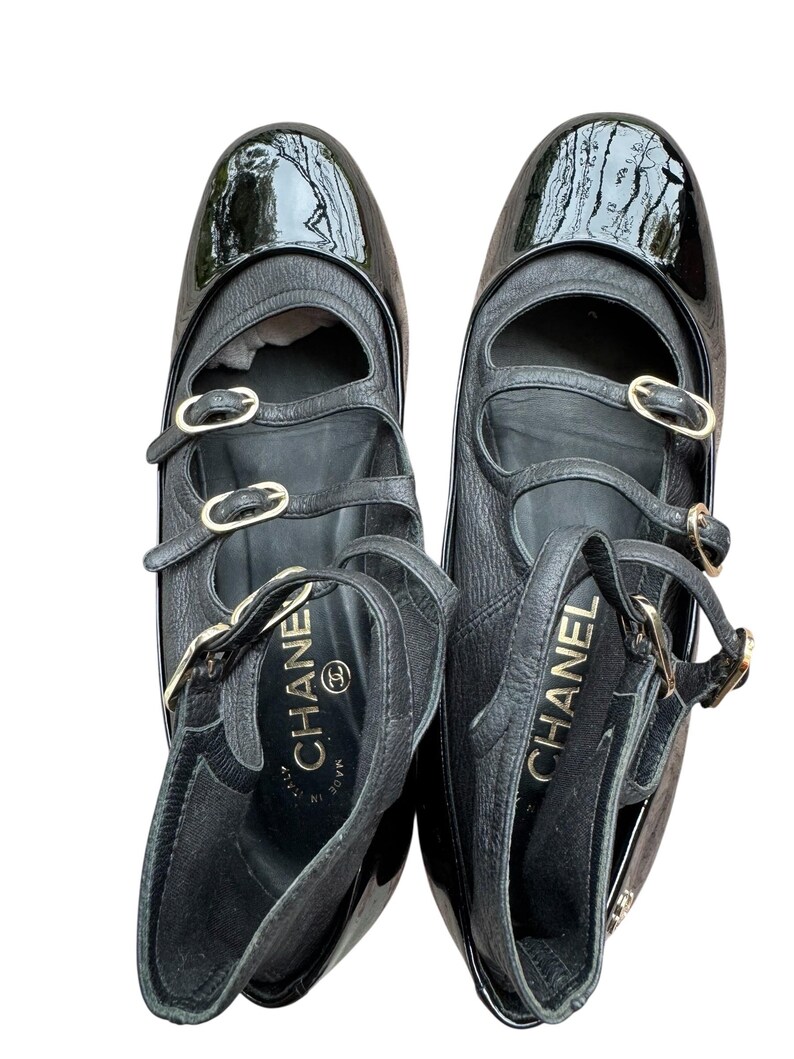 May include: Pair of black leather Mary Jane style shoes with patent leather toe caps. The shoes have multiple straps with gold-tone buckles and the brand name "CHANEL" is visible inside the shoe.