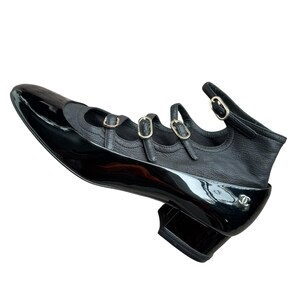 May include: Black leather and patent leather shoe with three buckle straps and a small heel. The shoe features a pointed toe and a logo on the side. The shoe is a stylish footwear option.
