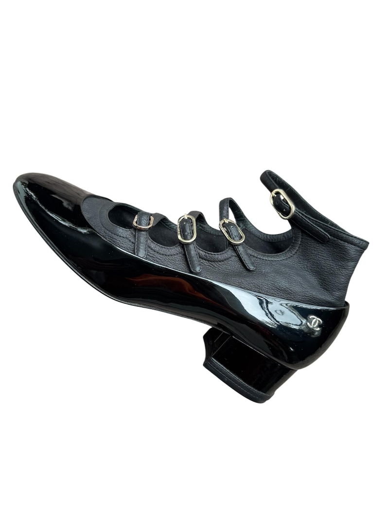 May include: Black leather and patent leather shoe with three buckle straps and a small heel. The shoe features a pointed toe and a logo on the side. The shoe is a stylish footwear option.