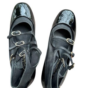 May include: Pair of black leather ballet flats with patent leather toe caps. The flats feature multiple straps with gold-tone buckles and the brand name embossed on the side. The shoes have a classic, elegant design.