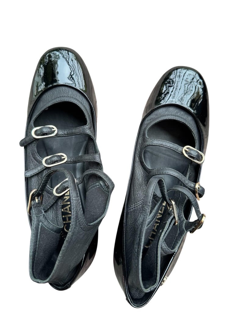 May include: Pair of black leather ballet flats with patent leather toe caps. The flats feature multiple straps with gold-tone buckles and the brand name embossed on the side. The shoes have a classic, elegant design.