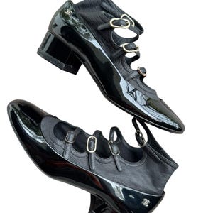 May include: Pair of black leather shoes with a glossy patent finish. The shoes feature multiple straps with gold-tone buckles and a low block heel. The design includes a rounded toe and a small logo detail.