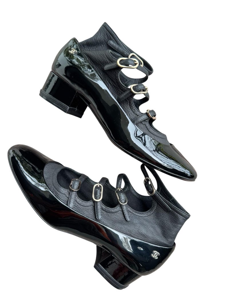 May include: Pair of black leather shoes with a glossy patent finish. The shoes feature multiple straps with gold-tone buckles and a low block heel. The design includes a rounded toe and a small logo detail.