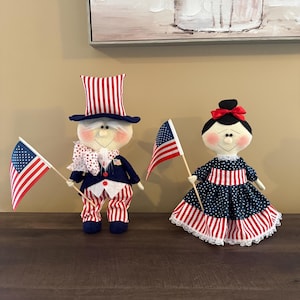 May include: Two patriotic dolls, one dressed as Uncle Sam with a red, white, and blue outfit and top hat, and the other in a blue and white dress. Both dolls hold miniature American flags, perfect for Independence Day.