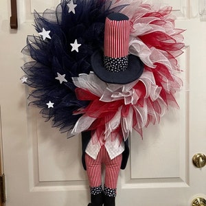 May include: A red, white and blue wreath with a fabric Uncle Sam figure in the centre. The wreath is made of mesh and has white stars on the blue mesh. The Uncle Sam figure is wearing a blue top hat with white stars, a red, white and blue striped waistcoat, and red, white and blue striped trousers.