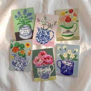 Puede incluir: Una colección de seis láminas rectangulares con ilustraciones de diversas flores en jarras y tazas de cerámica decorativas. Los diseños incluyen nomeolvides azules, flores rosas, amapolas rojas, capuchinas naranjas, peonías rosas y prímulas blancas.
