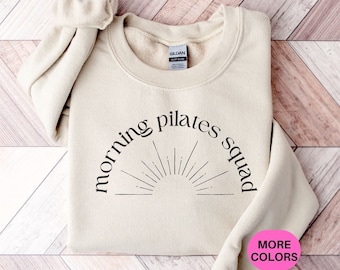 May include: A beige sweatshirt with the text "morning pilates squad" in black lettering and a sun graphic. The text is in an arc shape with the sun graphic in the center. The sweatshirt is on a wood background.
